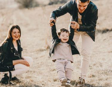 dad throws toddler while holding hands as mom watches and laughs